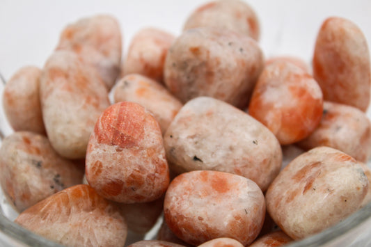 A collection of polished sunstone tumbles in various shades of red and orange, contained in a clear glass bowl.