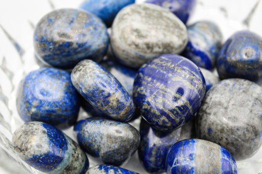 A collection of polished Lapis Lazuli stones with varying shades of blue and gold pyrite flecks, displayed in a clear bowl.