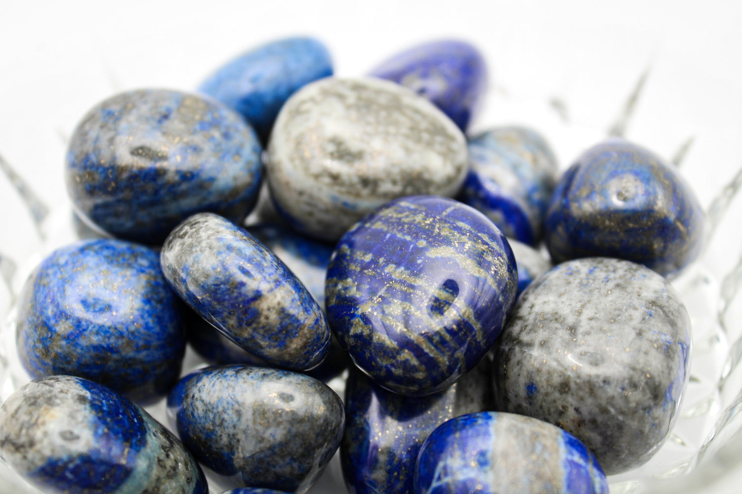 A collection of polished Lapis Lazuli stones with varying shades of blue and gold pyrite flecks, displayed in a clear bowl.